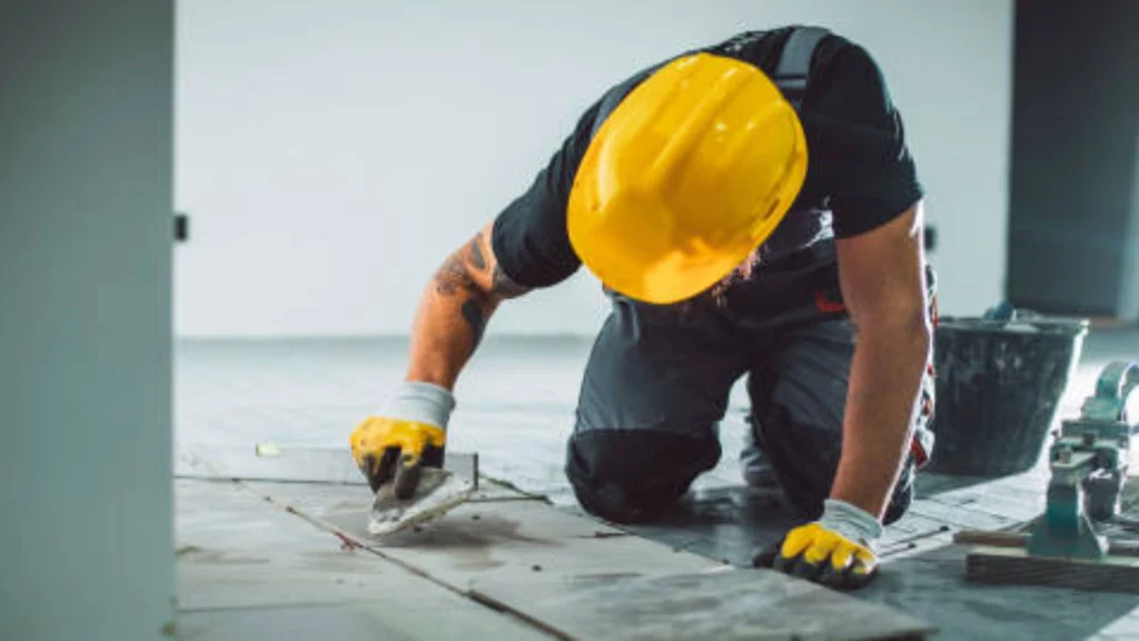 Construction worker installing floor tiles, wearing safety gear, using trowel tool. -bathroom remodel bellevue wa